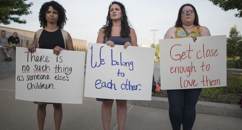 Three young women hold up signs reading "there is no such thing as somone else's children," "we belong to each other," and "get close enought to love them."