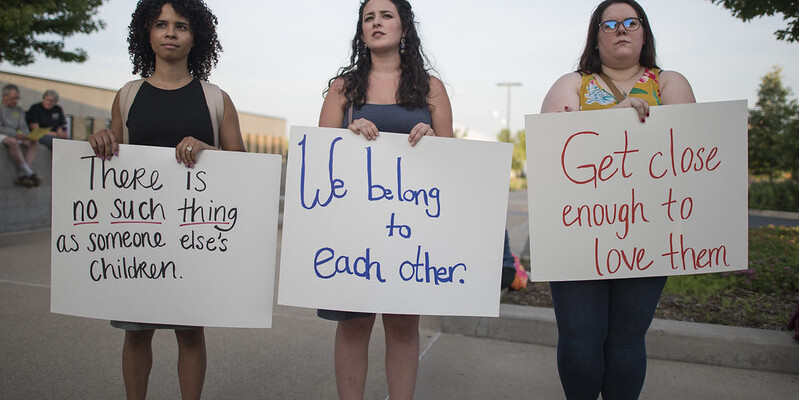 Three young women hold up signs reading "there is no such thing as somone else's children," "we belong to each other," and "get close enought to love them."