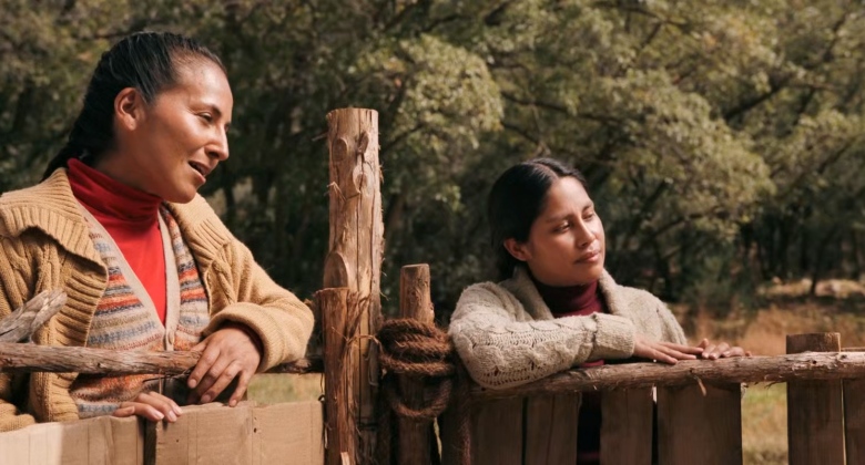 Two women leaning over fence as one woman sings in Quechua.